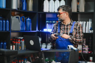 Araba parçası tutan mavi tulumlu bir satıcı ve oto yedek parça mağazasında dizüstü bilgisayarına bakıyor.