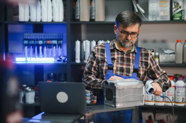 Kıdemli satıcı araba aküsünü bir oto yedek parça mağazasında barkod tarayıcısıyla tararken envanter yönetimi için dizüstü bilgisayar kullanıyor.