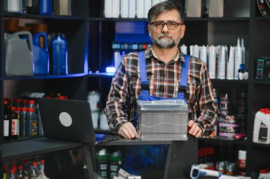 Tamirci, bir otomobil yedek parça mağazasında yeni bir akü gösteriyor. Etrafı ürünlerle dolu.