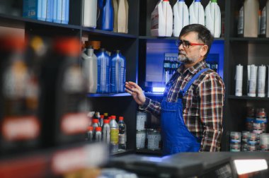 Oto yedek parça mağazası satıcısı rafta motor yağını kontrol ediyor, müşterisi için doğru ürünü seçiyor.