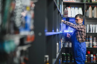 Kıdemli satıcı mavi tulumlar giyer. Parlak ışıklı bir oto yedek parça mağazasının rafındaki motor yağı şişelerini düzenler.