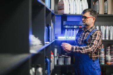 Gündüz vakti hareketli bir oto yedek parça mağazasında organize raflar arasında motor yağı şişesi arayan kıdemli bir satıcı.