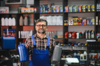 İki motor yağı şişesi tutan kıdemli satıcı, bir oto yedek parça mağazasında çalışıyor, raflar dolusu ürünlerle dolu.