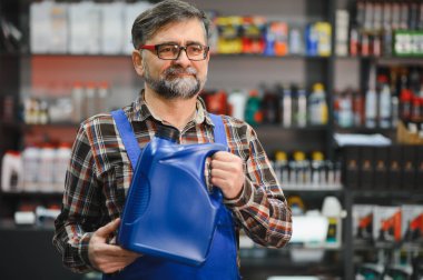 Bir oto yedek parça mağazasında müşterilere yardım ederken elinde büyük mavi bir şişe motor yağı tutan kıdemli satıcı, otomotiv uzmanlığı sergiliyor.