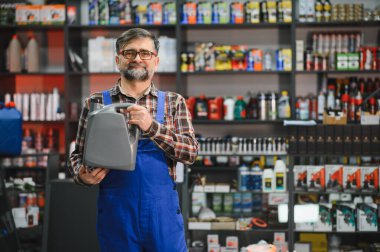 Mavi tulumlu satıcı bir oto yedek parça mağazasında gri bir motor yağı kutusu tutuyor ve arka planda bir sürü ürün var.