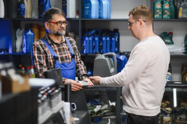 Tamirci motor yağı taşıyan müşteriden ödeme alıyor, malzeme dolu otomobil parçaları mağazasında dostça sohbet ediyor.