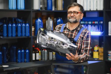 Bir oto yedek parça mağazasında çalışan kıdemli satıcı, elinde yeni bir araba farı tutarken müşterilerle kaliteli yedek parçalar hakkında konuşuyor.