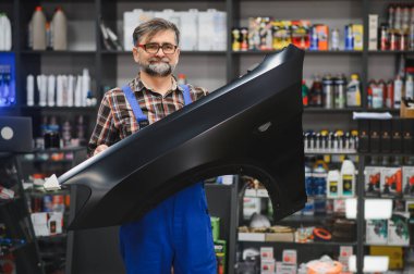 Oto yedek parça dükkanında siyah bir araba çamurluğu tutan kıdemli bir tamirci, arka planda çeşitli araba bakım ürünleriyle dolu raflar.