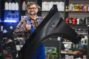 Tamirci, otomotiv ürünleriyle dolu bir oto yedek parça mağazasında yeni bir siyah araba tamponu sunarken,