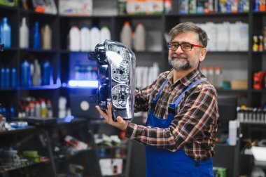 Kıdemli satıcı bir oto yedek parça mağazasında yeni bir far tutuyor. Etrafı ürünlerle dolu.