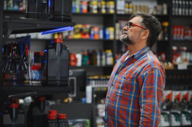 Bir oto yedek parça mağazasında motor yağı seçen kıdemli tamirci, ürünlerle dolu raflara bakıyor.