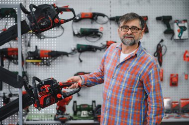 Bir hırdavatçıda testereyi sunan kıdemli bir satıcı, çeşitli elektrikli aletlerle çevrili, müşterilere profesyonel danışmanlık ve yardım sunuyor.