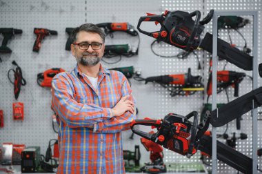 Çeşitli elektrikli testereler ve elektrikli testerelerle çevrili bir hırdavatçıda çapraz kollarla poz veren kendinden emin bir satıcı, uzmanlığını ve mağazanın ürün aralığını gösteriyor.
