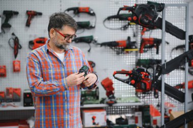 Sakallı olgun bir adam, vitrindeki çeşitli elektrikli aletlerle çevrili, hareketli bir hırdavatçıdan elektrikli matkap seçiyor.