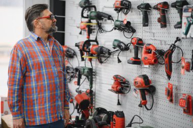 Sakallı, gözlüklü ve kırmızı ekose gömlekli olgun bir adam, bir hırdavatçıdan elektrikli elektrikli elektrikli aletler seçiyor.
