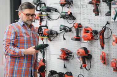 Sakallı müşteri bir hırdavatçıda matkap seçiyor ve duvarında bir sürü elektrikli alet asılı.