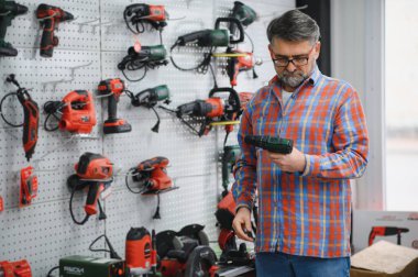Kıdemli adam bir hırdavatçıda kablosuz matkap seçiyor, duvarda çeşitli elektrikli aletler var.