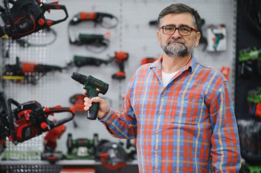 Bir hırdavatçıda kablosuz matkap tutan kıdemli satıcı, arka plandaki raflarda sergilenen elektrikli aletler