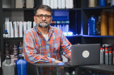 Sakallı satıcı oto yedek parça dükkanında dizüstü bilgisayar kullanıyor. Etrafı araba ürünleriyle dolu.