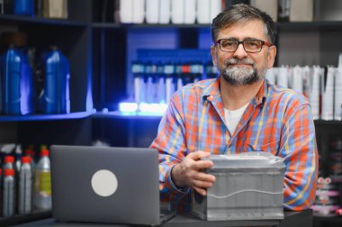 Satıcı ön planda dizüstü bilgisayarı olan bir araba aküsü tutuyor, bir oto yedek parça mağazasının içinde, raflarla dolu ürünlerle dolu.