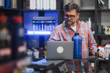 Satış elemanı dizüstü bilgisayardaki motor yağı bilgilerini kontrol ediyor ve oto yedek parça mağazasında barkod tarayıcısı kullanıyor.