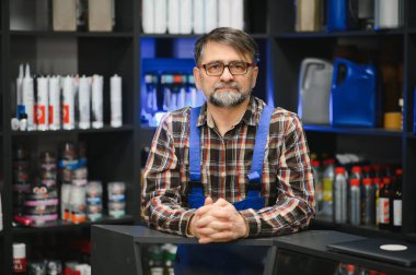 Bir oto yedek parça mağazasında elleri çapraz duran, raflarla çevrili kıdemli bir satıcı.