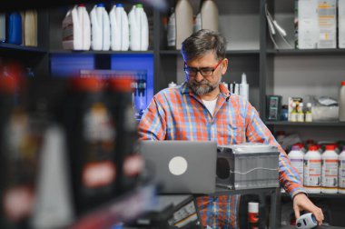 Bir araba aküsünü kontrol ederken dizüstü bilgisayar kullanan oto yedek parça mağazası satıcısı, etrafı ürünlerle dolu.