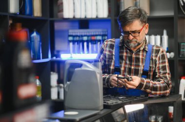 Bir oto yedek parça mağazasında mavi tulum giyen kıdemli satıcı, bir müşteriye satmadan önce işlevselliğini kontrol ediyor.