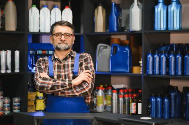 Otomobil yedek parça mağazasında duran kendinden emin bir satıcının portresi, çeşitli araba ürünleriyle dolu raflarla çevrili.