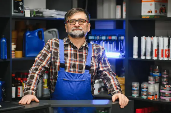 Araba bakım ürünleriyle dolu raflarla çevrili bir oto yedek parça mağazasında tezgaha yaslanmış kendinden emin bir satıcı.