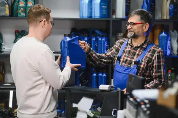 Tamircinin oto yedek parça mağazasında müşteriye bir kutu motor yağı vermesi, hareketli bir perakende ortamında dostane bir hizmet sergilemesi