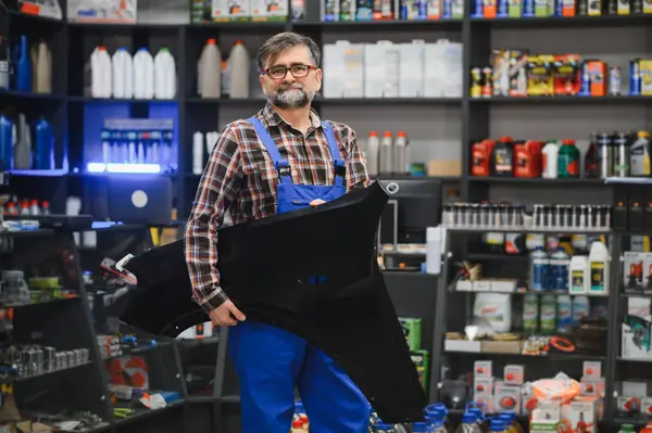 Satıcı elinde siyah plastik bir araba parçası tutuyor, muhtemelen çamurluk parçası, iyi stoklanmış bir oto yedek parça mağazasında.