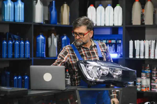 Dizüstü bilgisayarda çalışan kıdemli bir satıcı, otomobil yedek parça mağazasında araba farı bilgilerini kontrol ediyor, otomotiv malzemeleri raflarıyla çevrili.