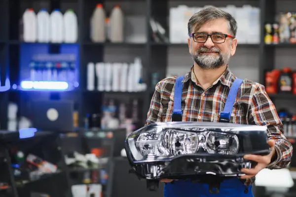 Gülümseyen olgun satıcı elinde yeni bir araba farı tutarken envanter ve aletlerle dolu bir oto yedek parça mağazasında ürünleri sergiliyor.