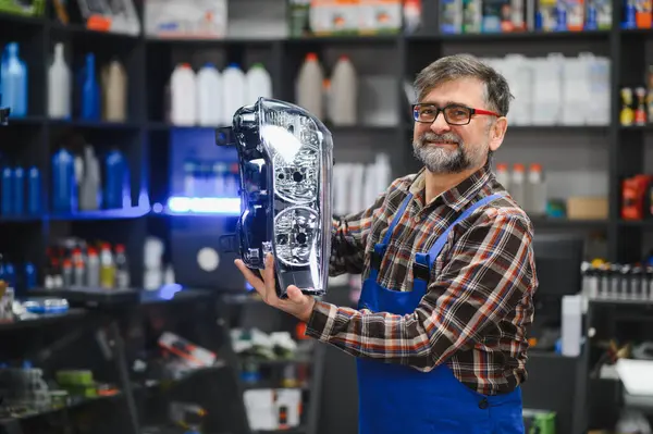 Kıdemli satıcı bir oto yedek parça mağazasında yeni bir far tutuyor. Etrafı ürünlerle dolu.