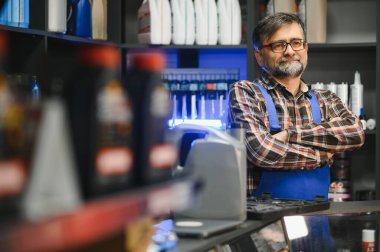 Oto yedek parça mağazasında çapraz kollarla poz veren kendinden emin olgun bir satıcı. Etrafı ürünlerle dolu.