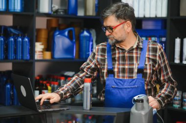 Bir oto yedek parça mağazasının tezgahında çalışan satıcı dizüstü bilgisayar kullanıyor ve elinde bir barkod tarayıcısı var.