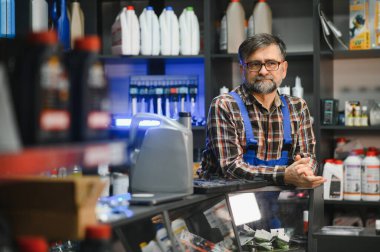 Sakallı ve gözlüklü kıdemli satıcı çeşitli otomotiv ürünleriyle dolu bir oto yedek parça mağazasında müşterileri sabırla bekliyor.