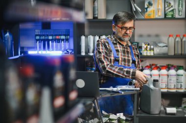 Satış elemanı mavi tulum giyiyor, oto yedek parça mağazasında müşterilere yardım ederken motor yağı şişesi tutuyor, otomotiv uzmanlığı sergiliyor.