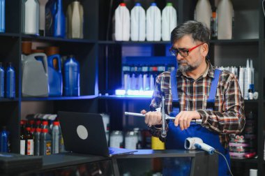 Kıdemli satıcı mavi üniformalı bir araba parçası tutuyor ve bir oto yedek parça dükkanında bilgisayarına bakıyor.