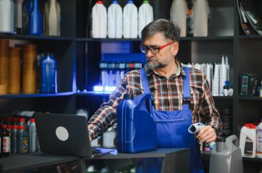 Barkod tarayıcı ve dizüstü bilgisayar kullanan kıdemli bir satıcı, otomobil yağı ürünleri deposundaki motor yağı bilgilerini doğruluyor, araba bakım ürünleriyle dolu raflarla çevrili.