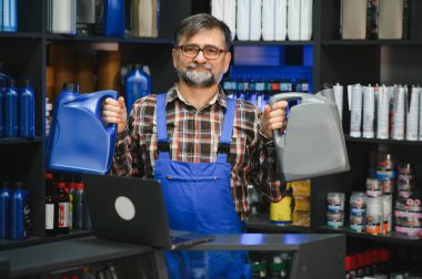 Bir oto yedek parça mağazasında çeşitli motor yağlarını sunarken özenle bir dizüstü bilgisayar üzerinde çalışan kıdemli satıcı, uzman tavsiyesi ile müşterilere yardımcı oluyor.