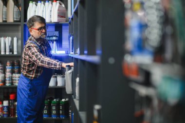 Kıdemli satıcı mavi tulumlar giyip oto yedek parça mağazasındaki bir rafta motor yağı şişelerini düzenliyor.