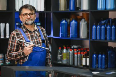 Araba yedek parça satıcısı elinde bir ingiliz anahtarı tutuyor. Etrafı araba bakım ürünleriyle dolu raflarla çevrili.