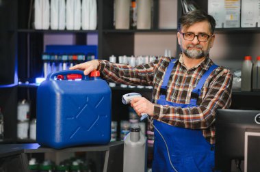 Kıdemli satıcı, oto yedek parça deposunda barkod tarayıcısıyla büyük mavi bidon yağ taraması yapıyor.