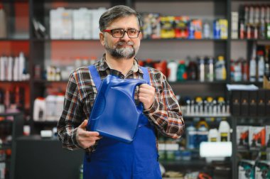 Bir oto yedek parça mağazasında büyük mavi bir teneke motor yağı tutan kıdemli bir satıcı, arka planda bir sürü ürünle dolu raflar