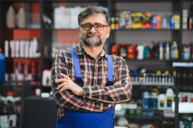 Olgun bir satıcının portresi, çeşitli malzemelerle çevrili iyi stoklanmış bir oto yedek parça mağazasında dikilirken güvenle kollarını kavuşturuyor.