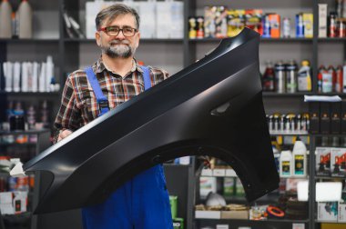 Bir oto yedek parça dükkanında siyah bir araba çamurluğu tutan tamirci, arka planda çeşitli araba bakım ürünleriyle dolu raflar