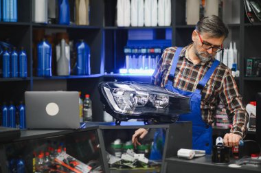 Araba yedek parça satıcısı mavi tulumlu, elinde araba farıyla yağ filtresi seçiyor.