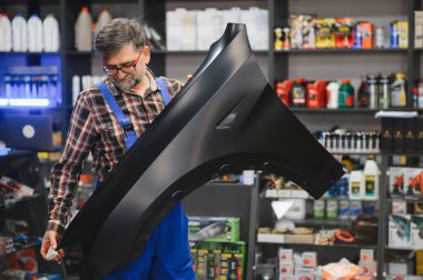 Tamirci, otomobil parçaları satan bir dükkanda içi ürünlerle dolu siyah bir araba tamponu tutuyor.